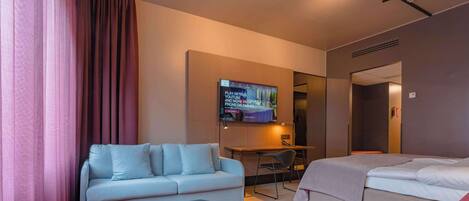 Ein Hotelzimmer mit einem hellblauen Sofa, einem pinkfarbenen Sessel, einem kleinen schwarzen Tisch und einem Bett mit grauer Decke.