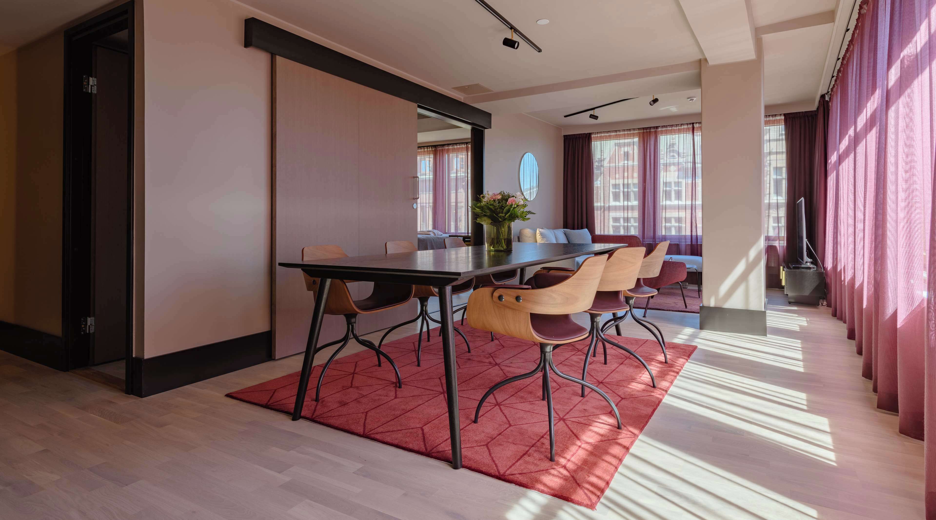 A modern hotel room with a dining area featuring a long table, multiple chairs, and a red patterned rug.