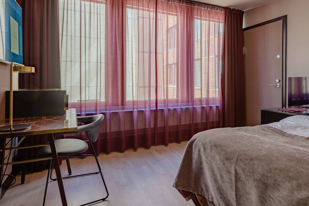 A hotel room with a bed, desk, chair, and a window with sheer curtains.