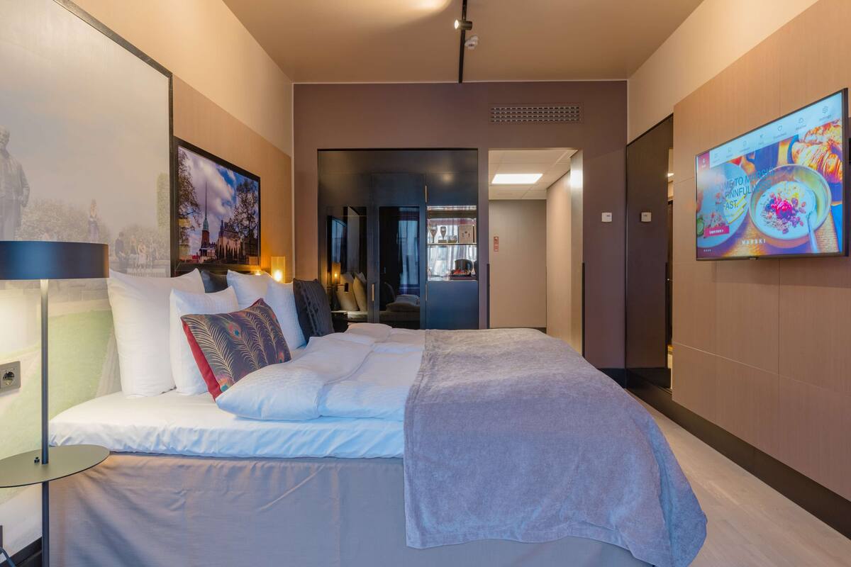 A modern hotel room with a large bed, a flat-screen TV mounted on the wall, and a view of the cityscape through the window.