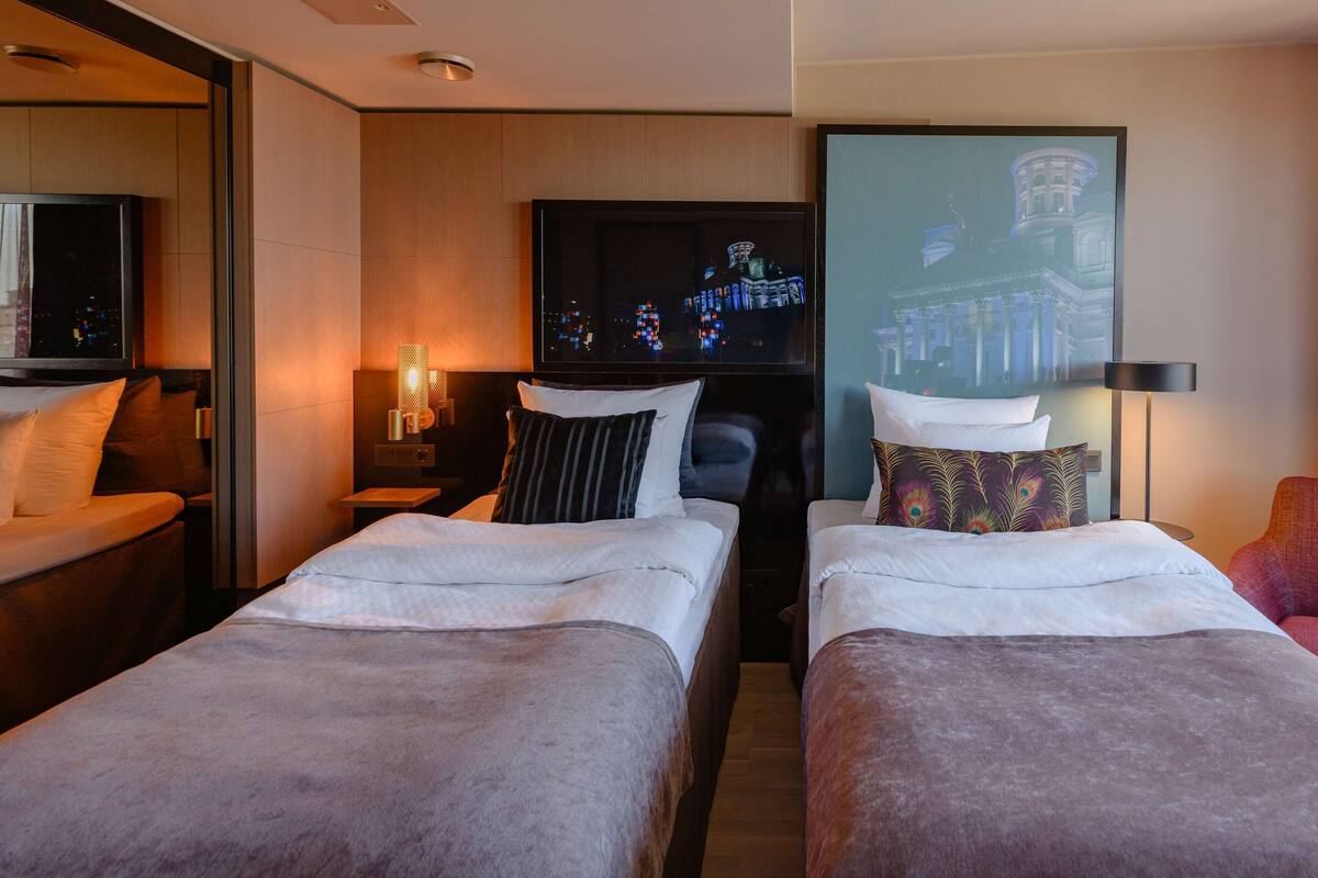 A hotel room with two single beds, a TV, a nightstand, a lamp, and a window with curtains.