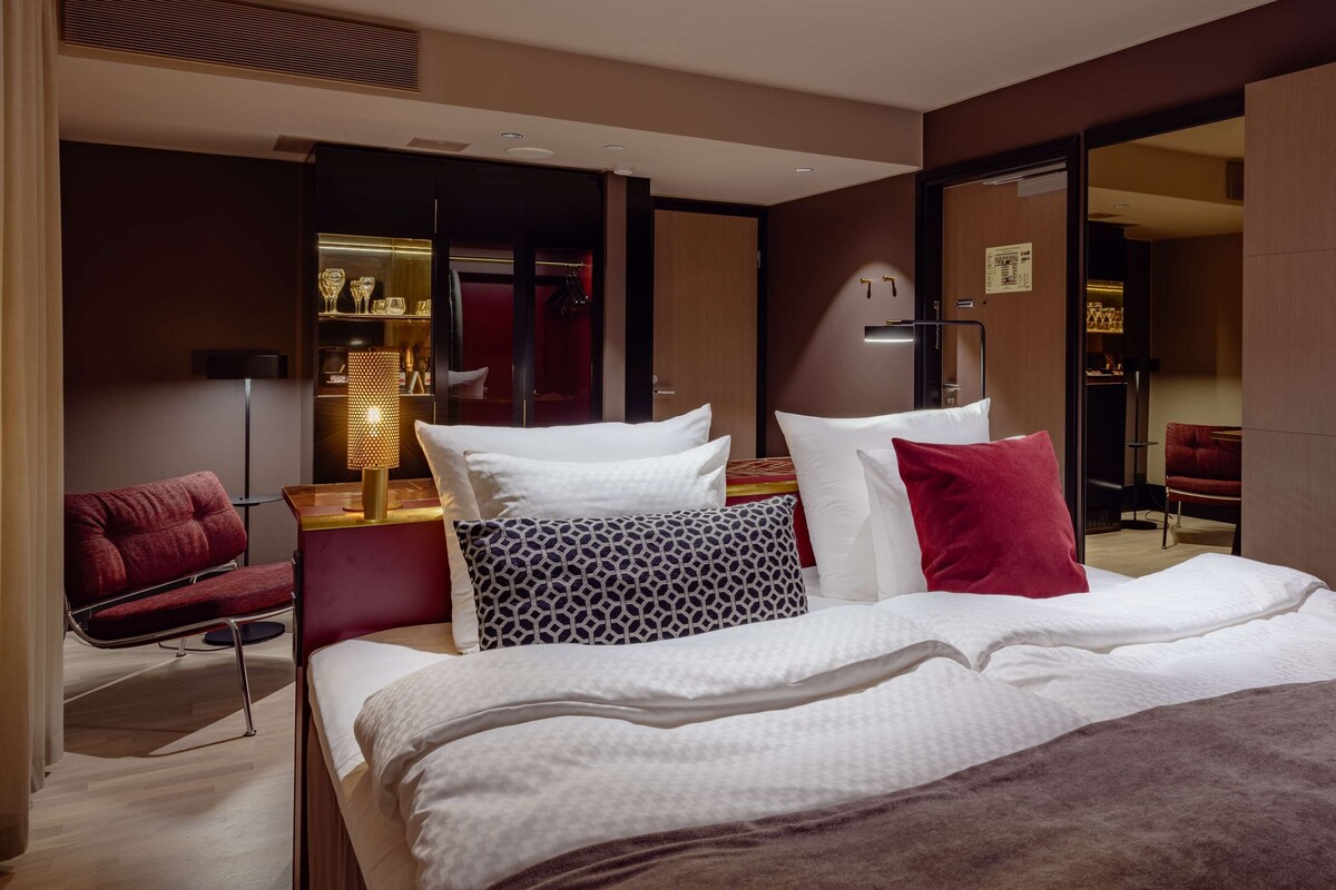 Habitación de hotel moderna con una cama grande, un sillón rojo, una mesita de noche con lámpara y un armario.