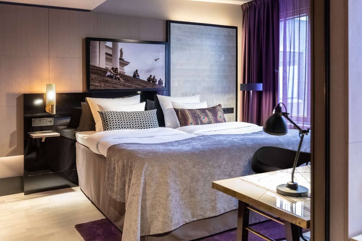 A modern hotel room with a large bed, a desk with a lamp, a TV mounted on the wall, and a window with curtains.