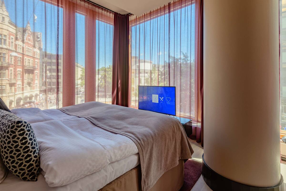 A hotel room with a bed, a TV, and a view of a cityscape through large windows.
