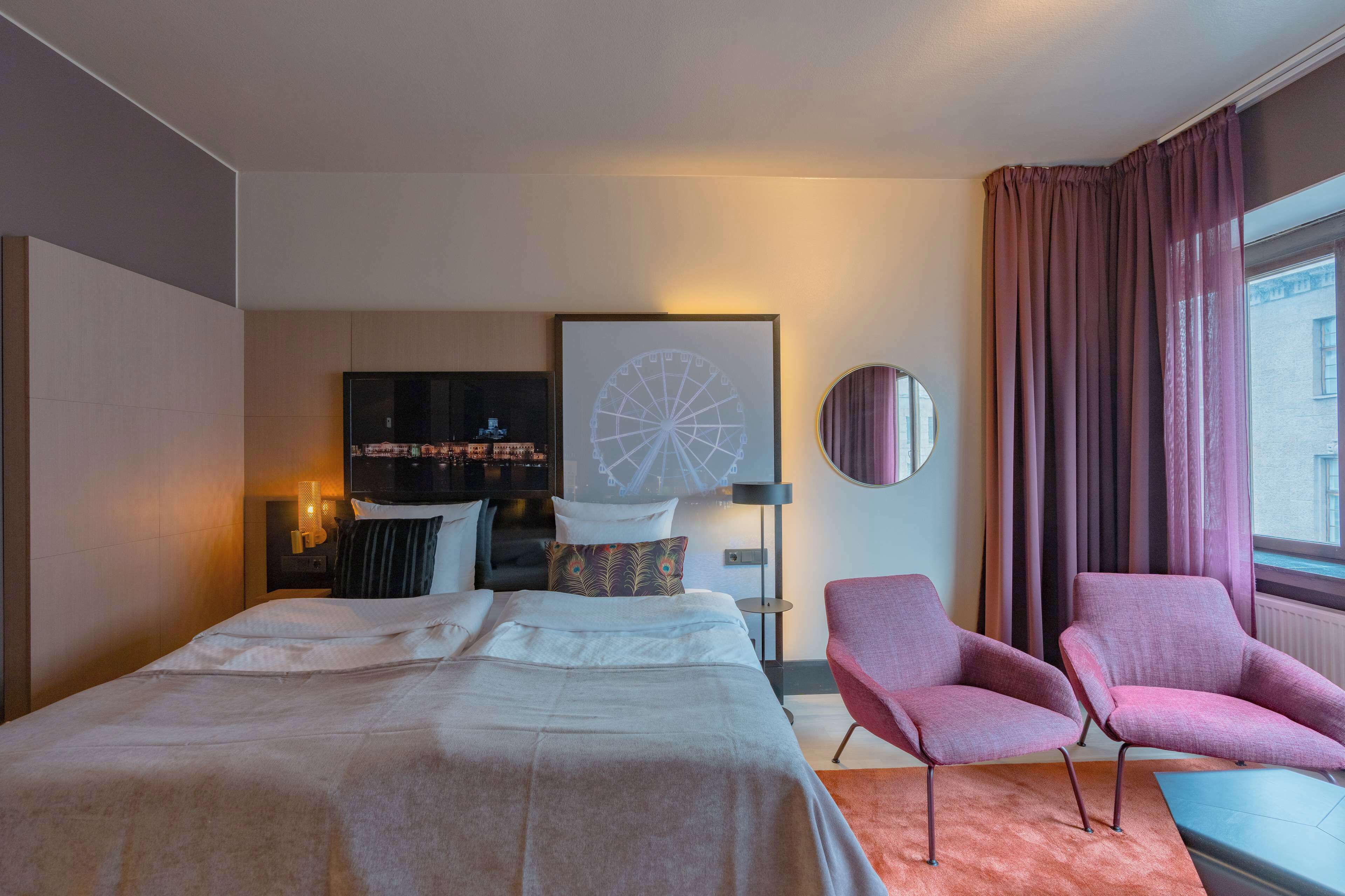 A modern hotel room with a large bed, a TV, a round mirror, and two pink armchairs.