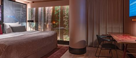Habitación de hotel moderna con una cama grande, un escritorio con sillas, un ventanal con vistas a un bosque y una pantalla de televisión mostrando la interfaz del hotel.