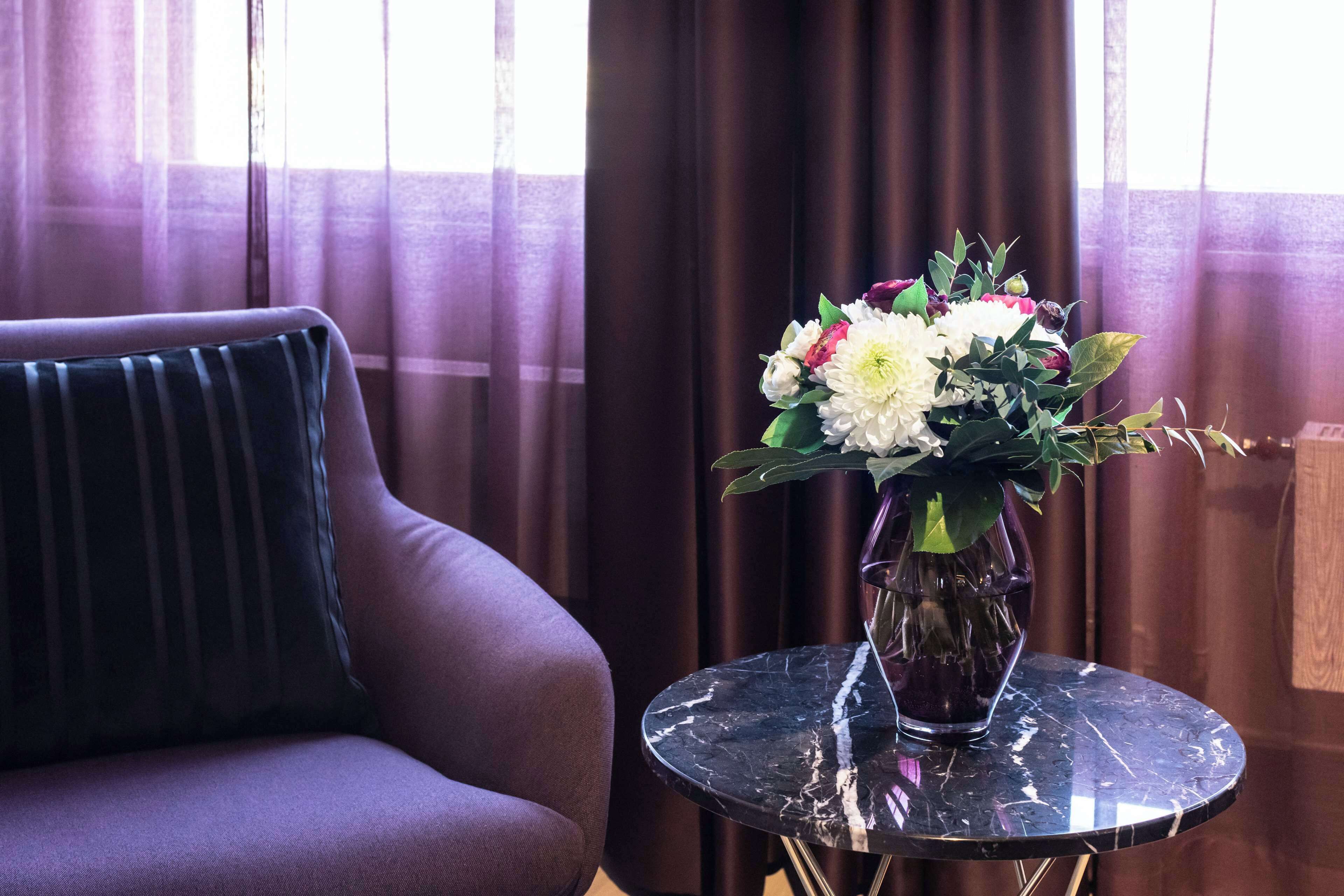 A room with a dark brown sofa, a small round table with a vase of flowers, and a window with sheer curtains.