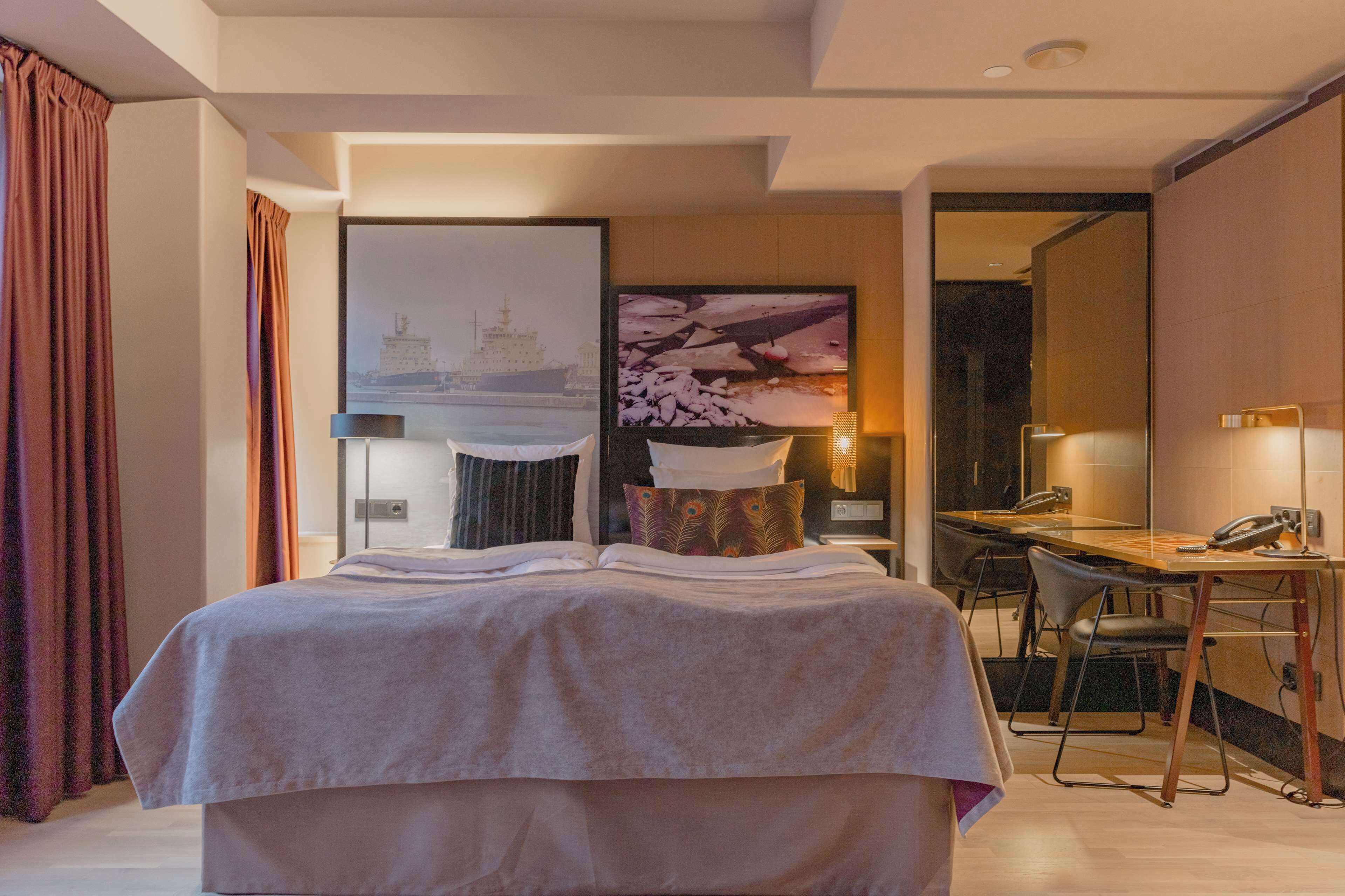 Ein modernes Hotelzimmer mit einem großen Bett, einem Schreibtisch und einem Stuhl. Ein Bild hängt an der Wand und ein Fenster mit Vorhängen ist vorhanden.