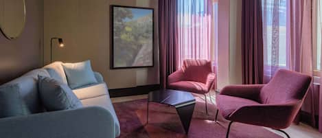 Una sala moderna con un sofá gris, un sillón rojo y una mesa de centro negra. Hay una foto grande con marco en la pared y una ventana con cortinas moradas.