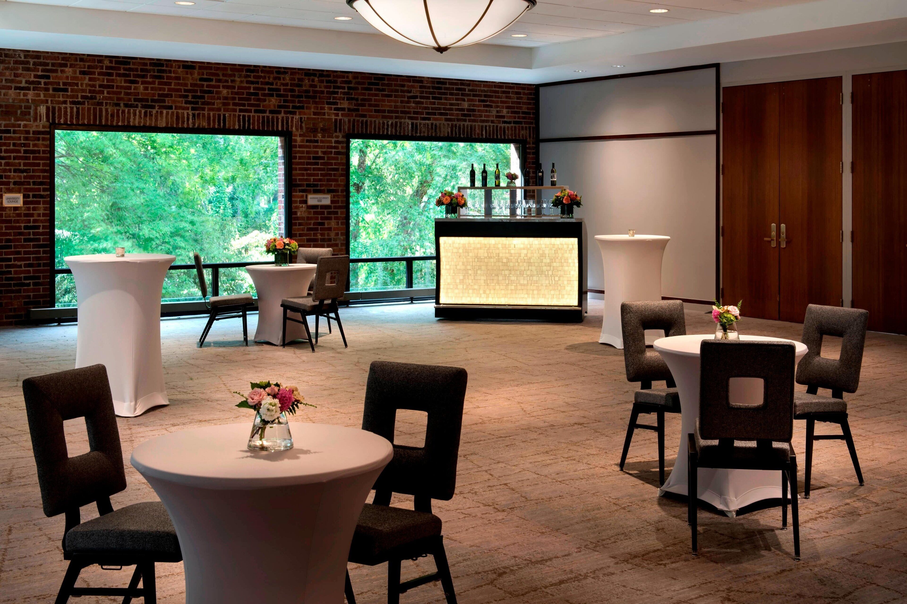 Une salle de conférence dotée de tables et de chaises rondes, un coin bar avec des bouteilles et des fleurs, et de grandes fenêtres offrant une vue sur la verdure.