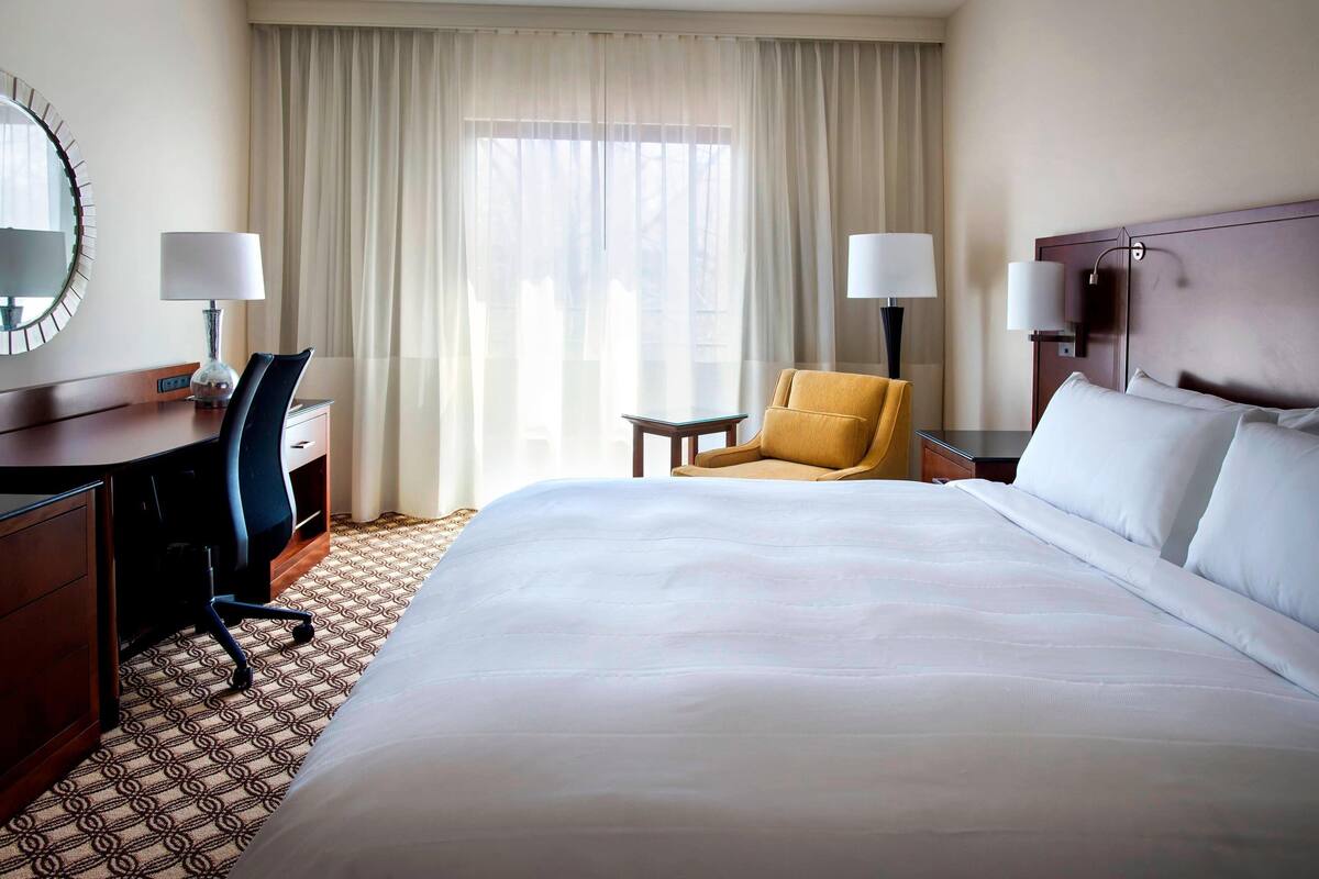 Ein Hotelzimmer mit einem großen Bett, zwei Stühlen, einem Schreibtisch, einer Lampe und einem Fenster mit Vorhängen.