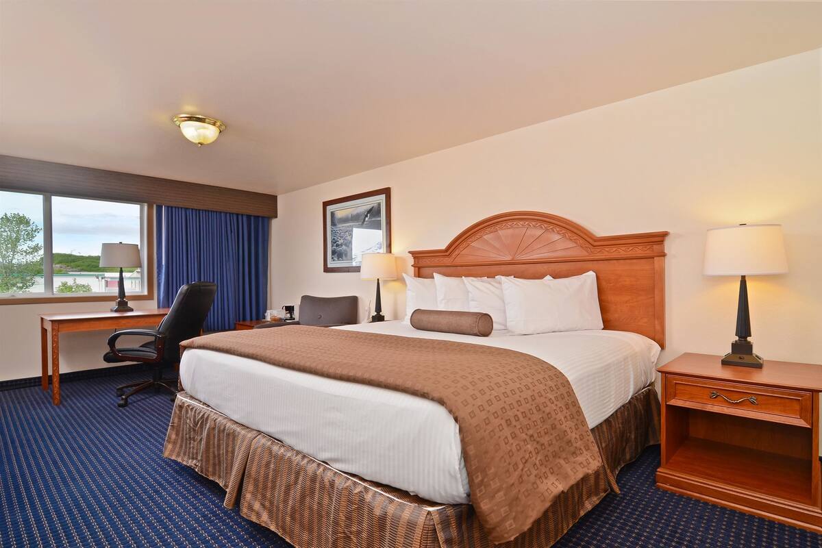 Habitación de hotel con una cama grande, un escritorio, una silla, dos lámparas de noche, una mesita de noche y una ventana con vistas al exterior.