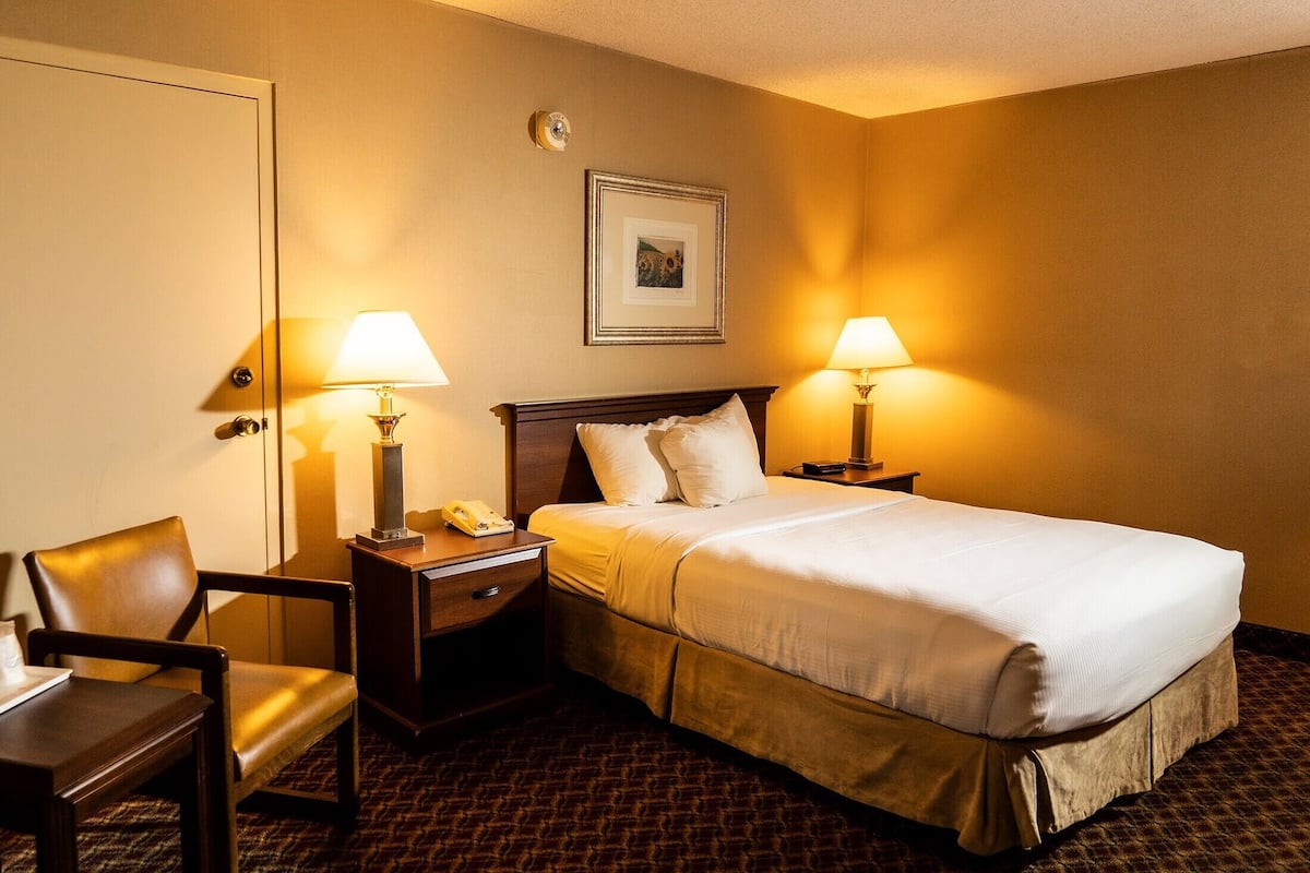 Een hotelkamer met een bed, twee bedlampen, een stoel, een nachtkastje en een ingelijst schilderij aan de muur.