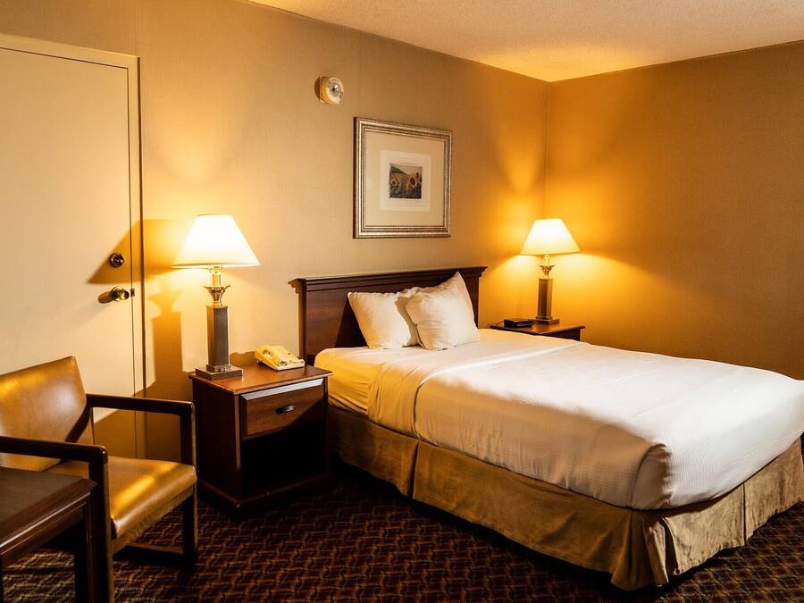 Habitación de hotel con cama, dos lámparas de noche, una silla, mesita de noche y un cuadro en la pared.