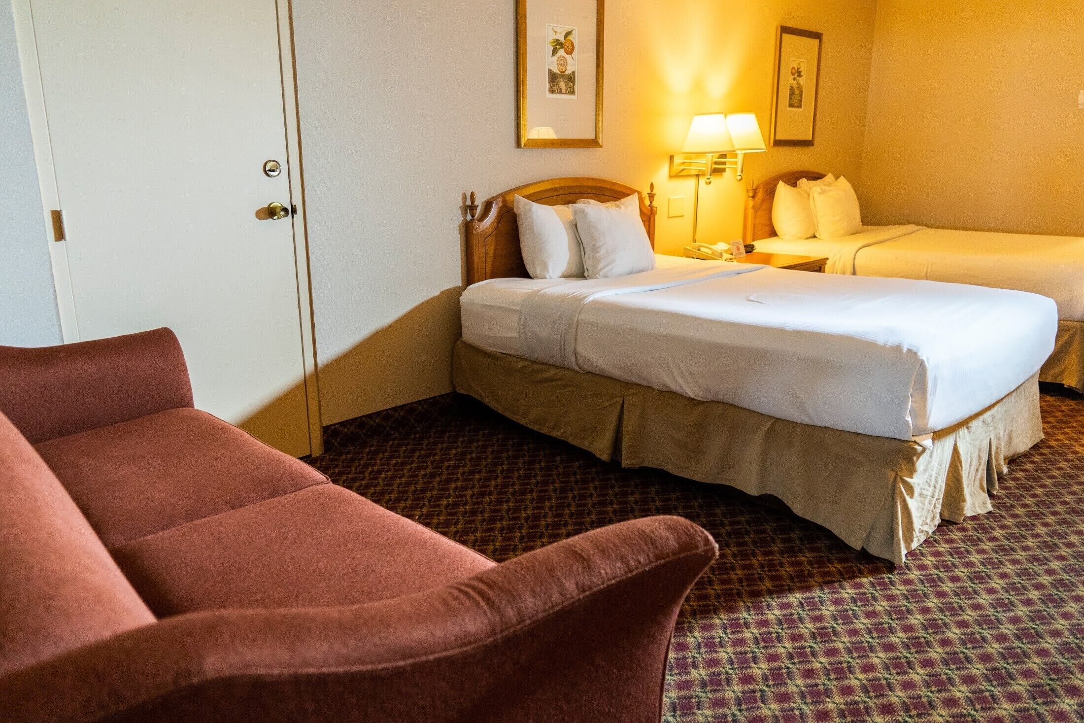 A hotel room with two beds, a wooden headboard, a red armchair, a patterned carpet, and a door.