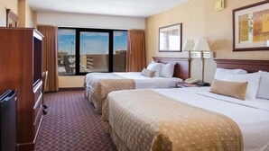 Ein Hotelzimmer mit zwei Betten, einem Schreibtisch, einem Stuhl, einem Fenster mit Blick auf die Stadt und einem gerahmten Bild an der Wand.