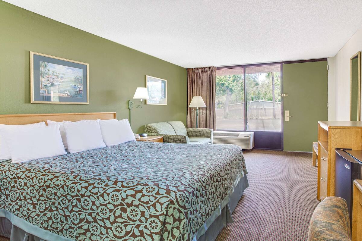Ein Hotelzimmer mit einem großen Bett, einem Schreibtisch, einem Stuhl, einer Lampe und Blick ins Freie.