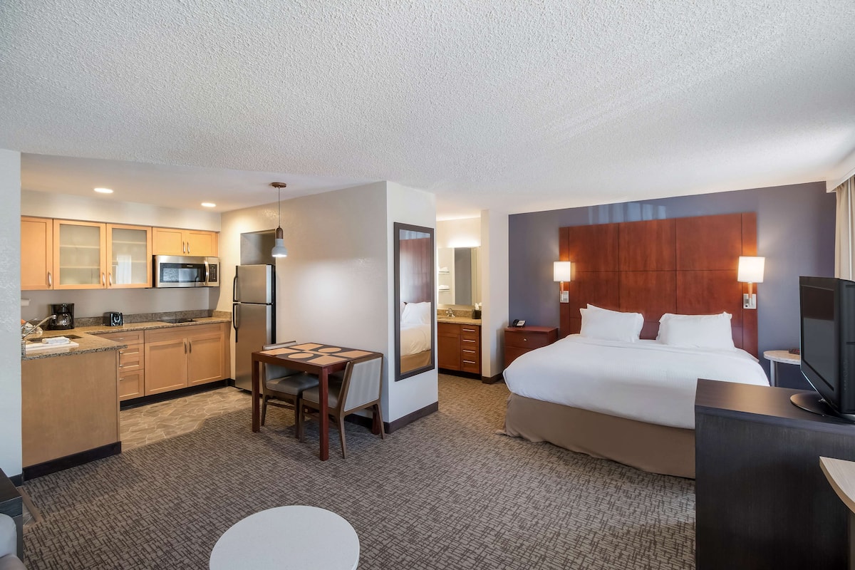 A hotel room with a bed, a desk, a chair, a small table, a television, and a kitchenette with a refrigerator, microwave, and coffee maker.