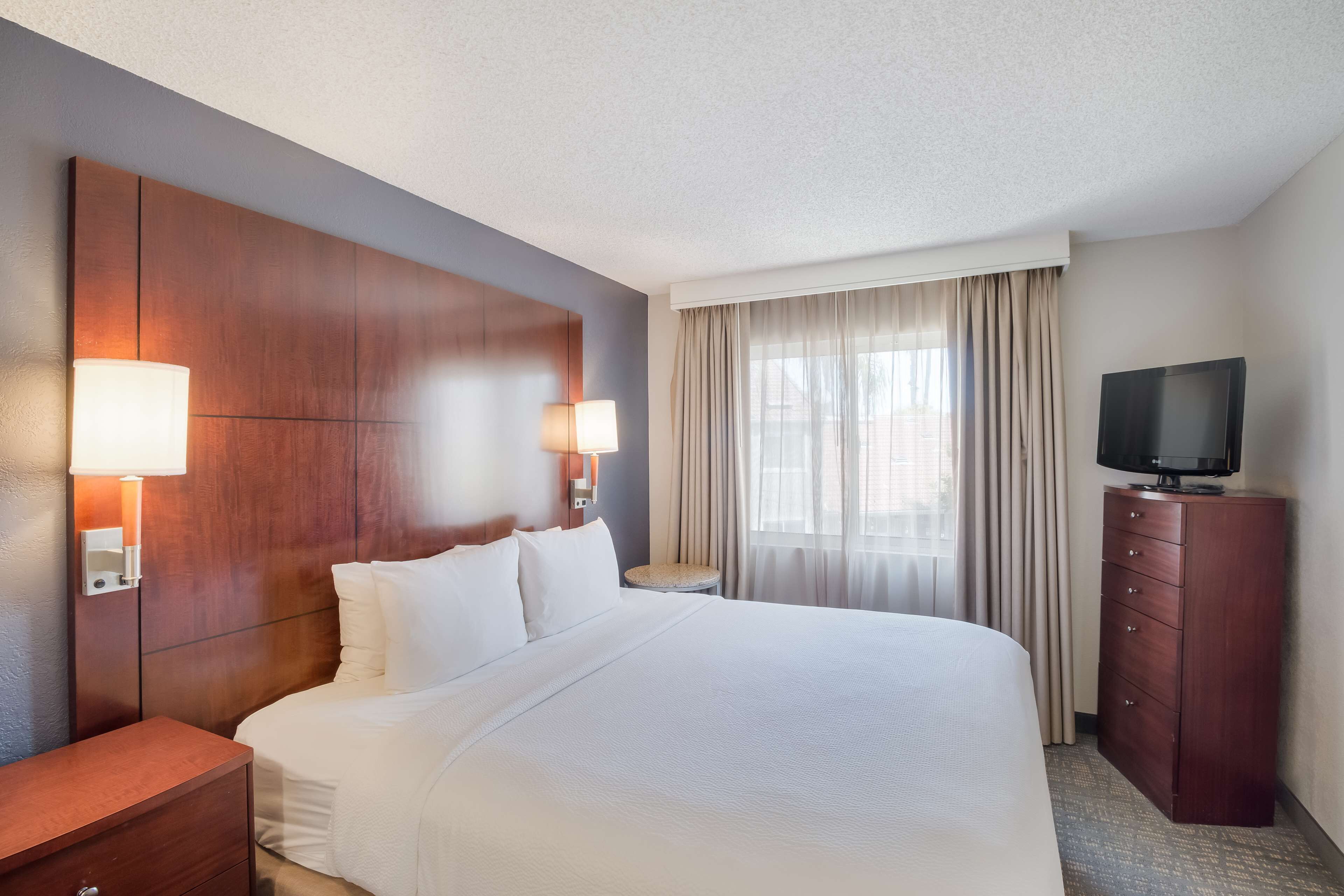 Habitación de hotel con una cama grande, dos lámparas de noche, un televisor sobre un mueble de madera y una ventana con cortinas.