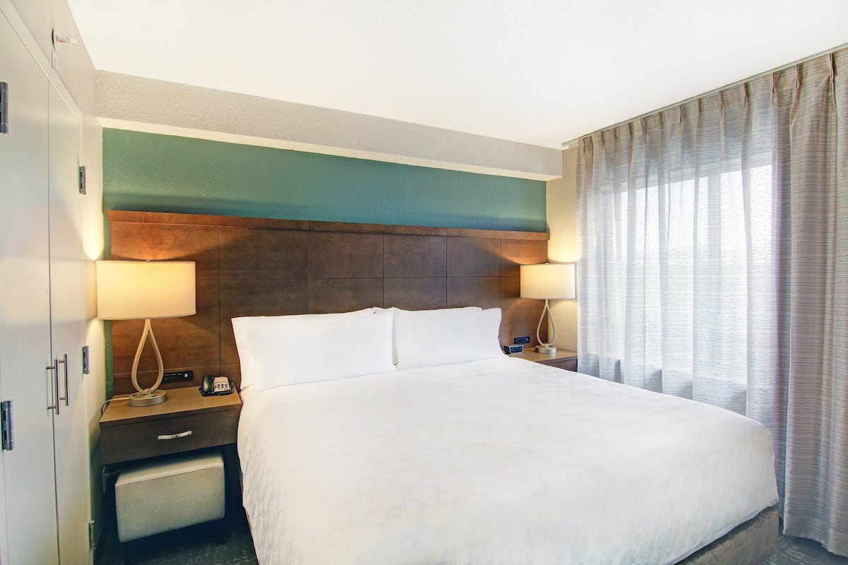 Une chambre d’hôtel avec un grand lit, deux tables de chevet avec des lampes, une table de nuit, une armoire et une fenêtre avec des rideaux.