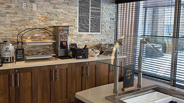 A kitchen area with a stone wall, a coffee machine, a food display, and a window with blinds. - Staybridge Suites Toronto - Vaughan South by IHG (Vaughan)