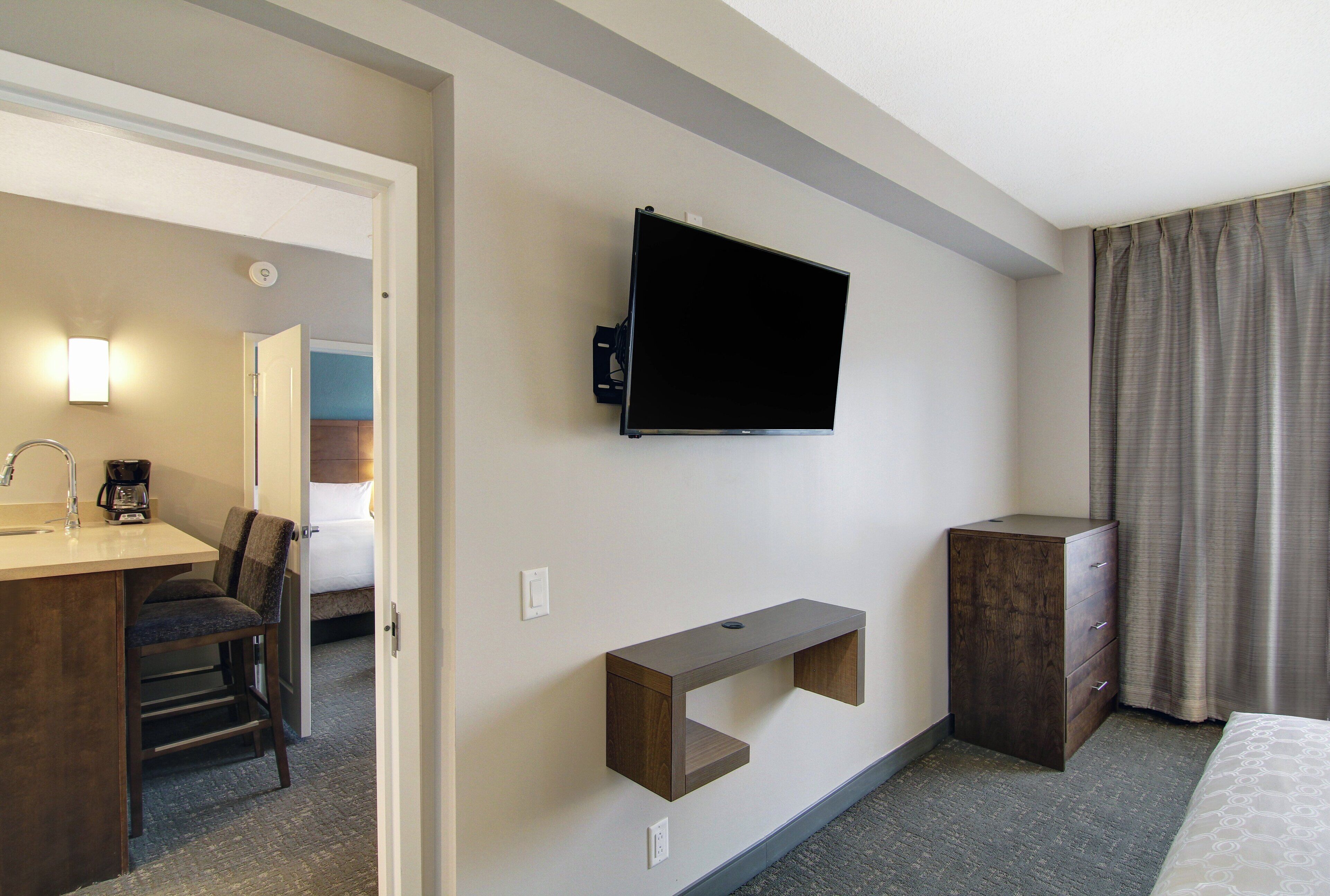 A hotel room with a flat-screen TV mounted on the wall, a desk with a chair, a small wooden shelf, a bedside cabinet, and a bed with a patterned bedspread.