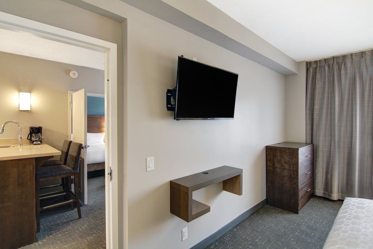 Une chambre d’hôtel comprenant un lit, un téléviseur fixé au mur, un petit bureau avec une chaise et une commode en bois.
