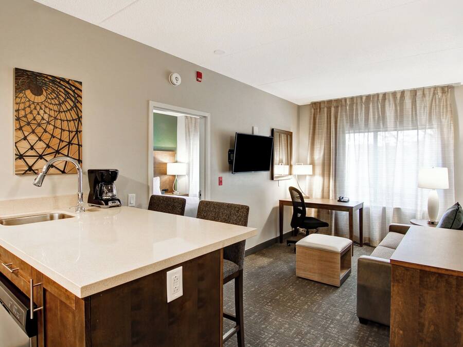 A hotel room with a kitchenette, a dining area, a living area with a sofa and armchair, a flat-screen TV mounted on the wall, and a large window with curtains.