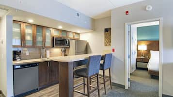 A hotel kitchen with a microwave, coffee maker, and a sink. There is a bar area with two chairs. The room has a door leading to a bedroom with a bed and a nightstand.