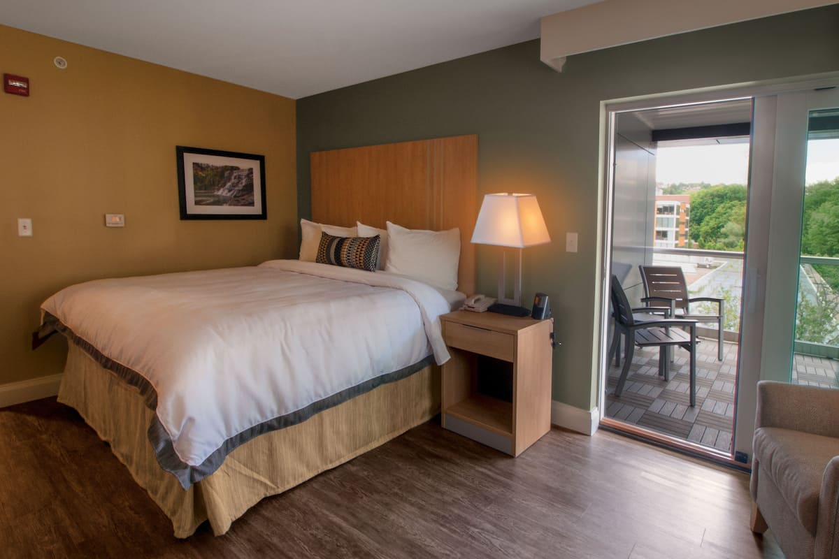 A hotel room with a large bed, a bedside table with a lamp, a chair, and a view of the outdoors through a sliding glass door.