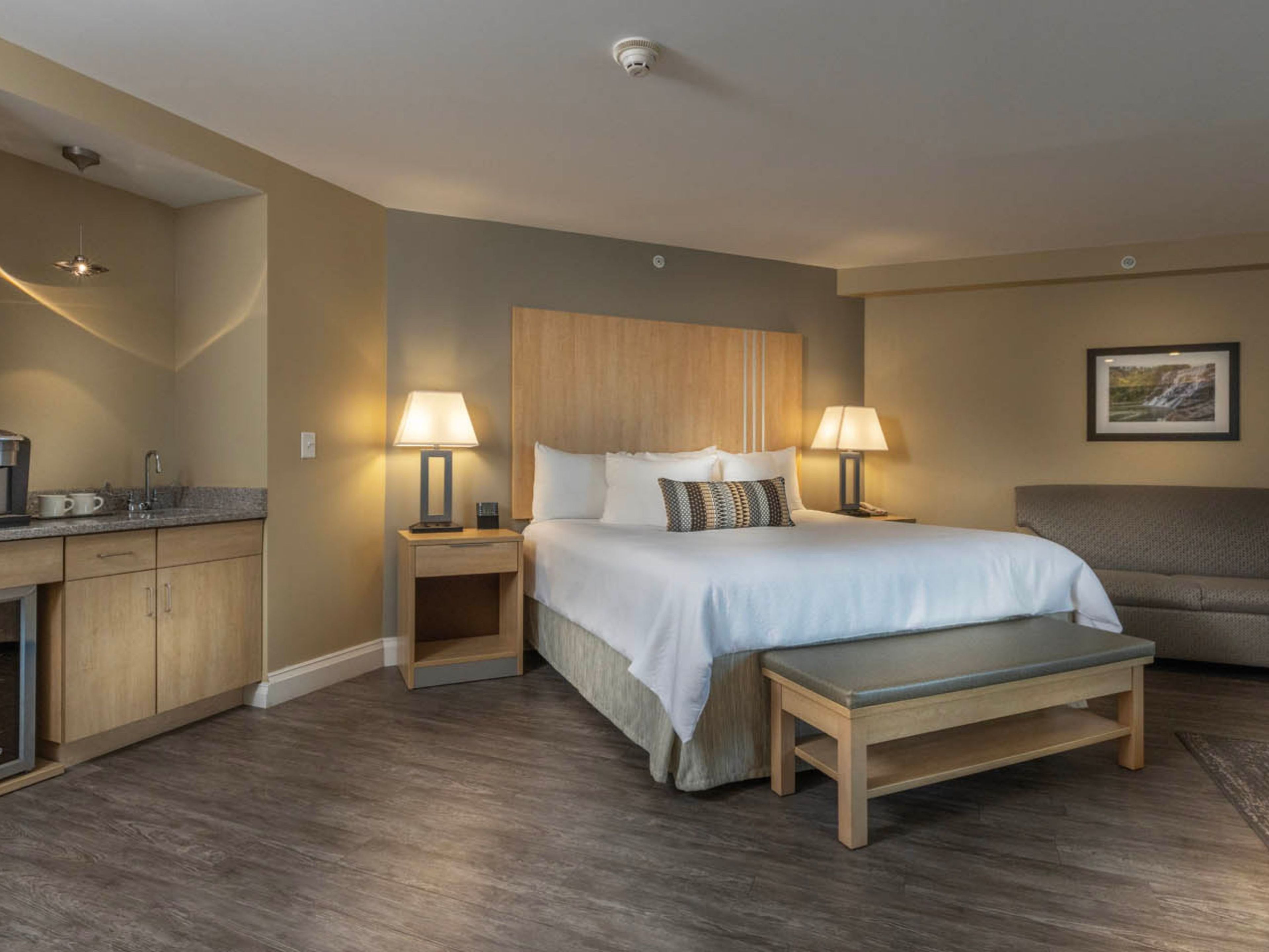 A hotel room with a bed, bedside tables, a bench, a sofa, a coffee machine, and a picture on the wall.