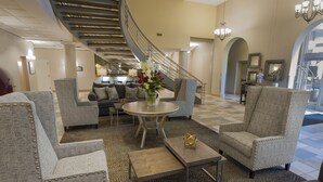 A spacious lobby with a central seating area featuring armchairs and sofas, a round coffee table, and a decorative vase. A staircase with a railing leads to an upper level. - Hotel Ithaca (Ithaca)