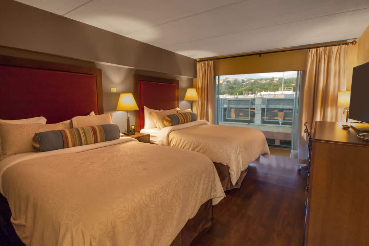 A hotel room with two beds, a wooden dresser, and a view of a cityscape through large windows.