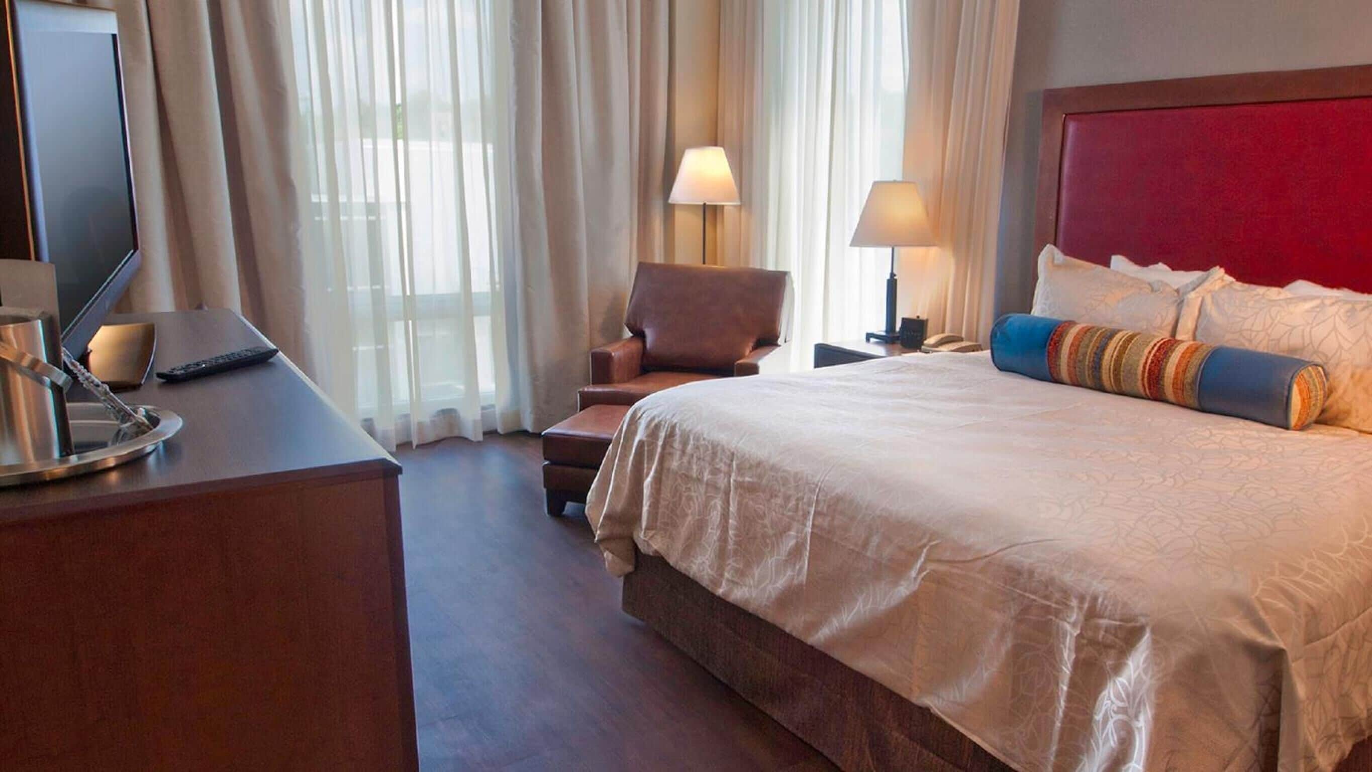 A hotel room with a large bed, a desk with a lamp, a chair, and a TV.