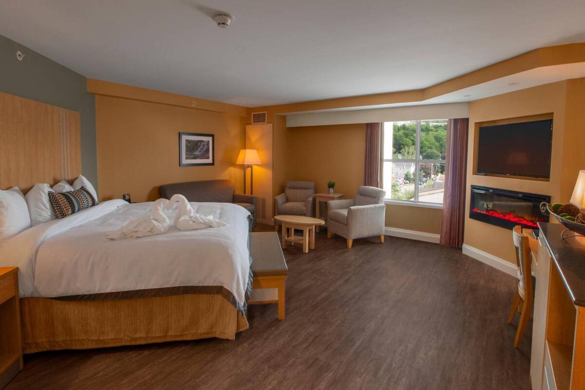 A hotel room with a large bed, a desk with a flat-screen TV, a fireplace, and a view of the outdoors.