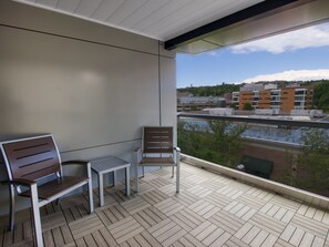 A balcony with two wooden chairs and a small table, overlooking a cityscape and a body of water.