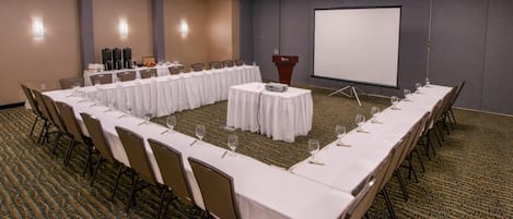 Un salón de conferencias con una larga mesa, sillas, un atril y una pantalla de proyector.