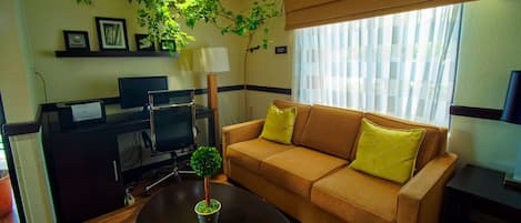 A cozy living room with a sofa, a coffee table, a desk with a computer, and a window with blinds.
