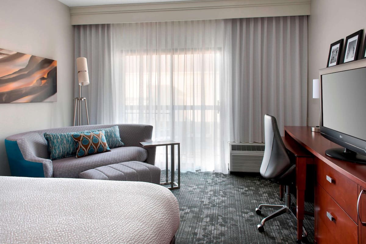 Een hotelkamer met een bed, een bank, een bureau, een stoel, een televisie en een groot raam met gordijnen.