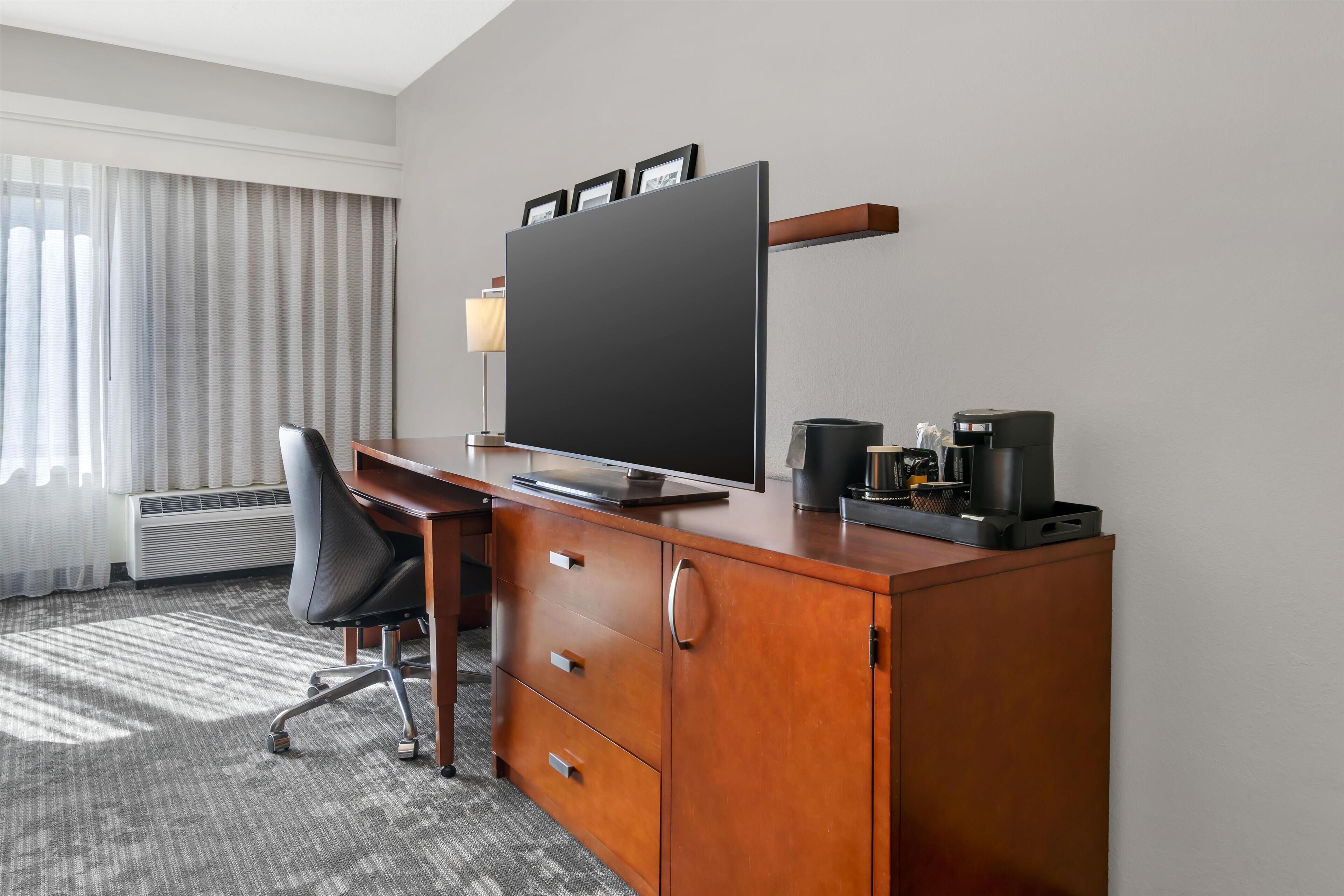 A hotel room with a wooden desk, a flat-screen TV, a coffee maker, and a chair.