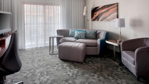 A hotel room with a grey sofa, a matching ottoman, a small side table, a TV, and a lamp.