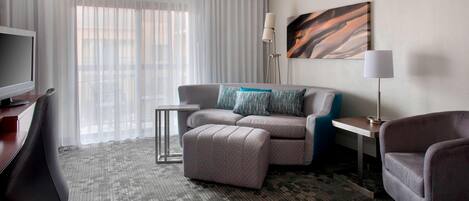 Una habitación de hotel con un sofá gris, un reposapiés a juego, una mesita auxiliar pequeña, un televisor y una lámpara.