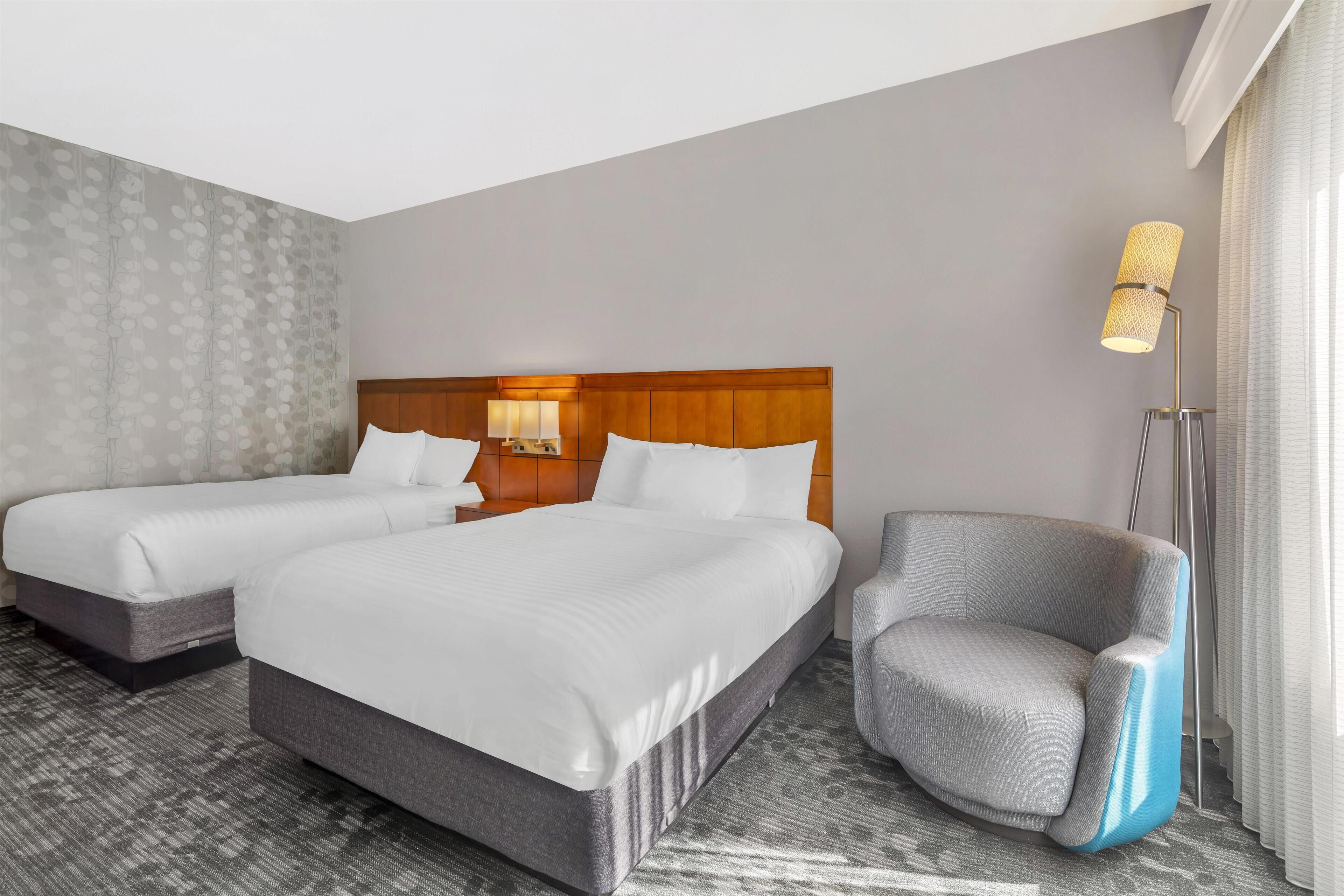 Quarto de hotel com duas camas, uma poltrona cinza, um abajur e uma parede estampada.