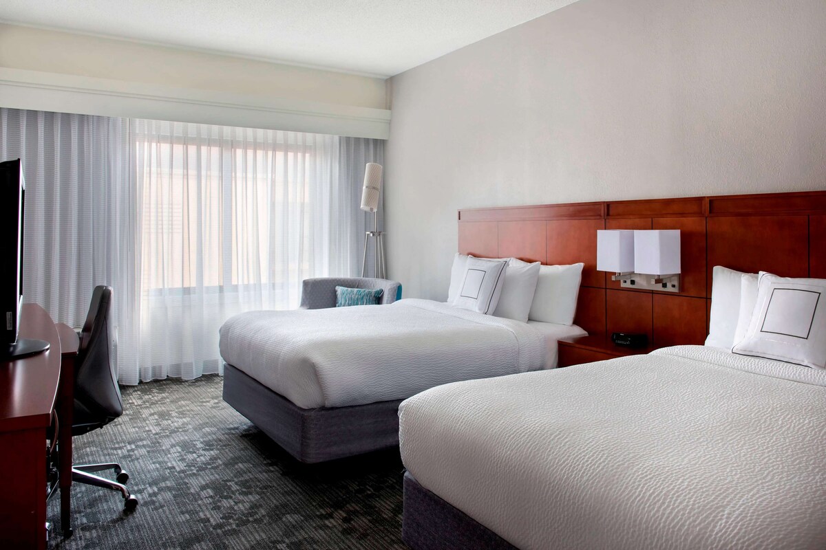A hotel room with two beds, a desk, a chair, a lamp, and a window with curtains.