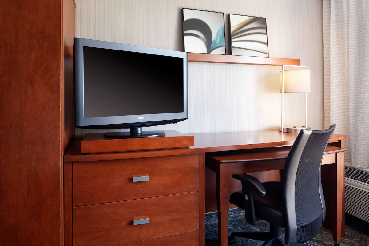 A hotel room with a wooden desk, a flat-screen TV, a chair, and a lamp.
