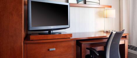 A hotel room with a wooden desk, a flat-screen TV, a chair, and a lamp.