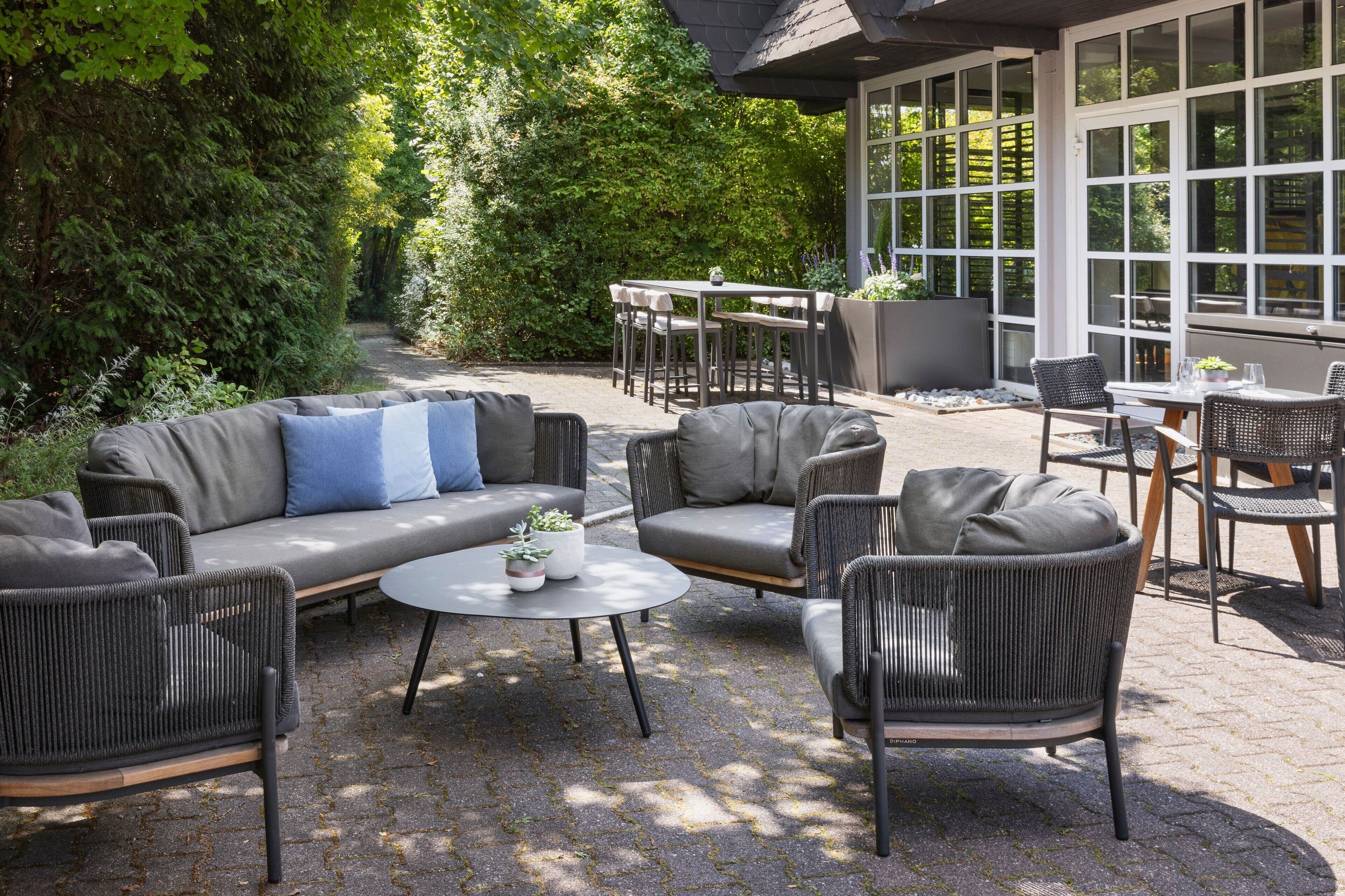 Un espace extérieur aménagé avec un canapé, des fauteuils et des tables, entouré de verdure et bordé d’un bâtiment doté de grandes fenêtres.