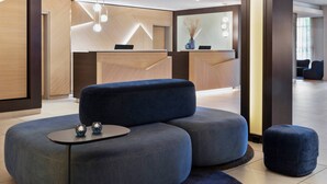 A modern reception area with a wooden reception desk, a blue seating area with ottomans, and a small table with decorative items. - Courtyard by Marriott Wiesbaden-Nordenstadt (Wiesbaden)