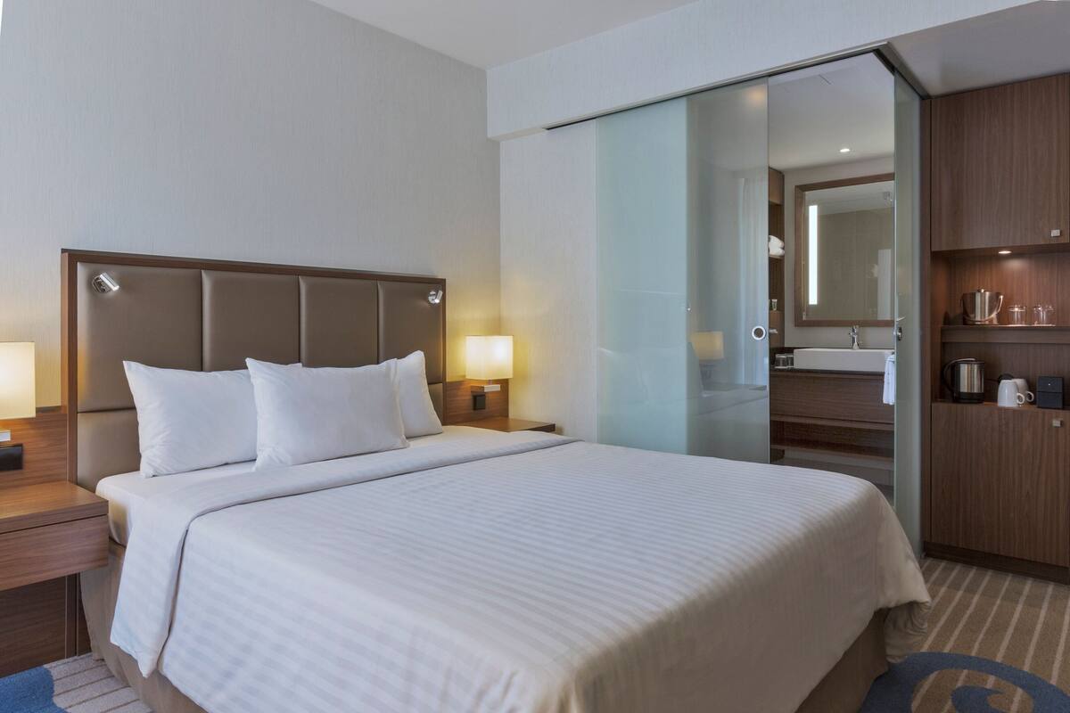 Ein Hotelzimmer mit einem großen Bett, einem Nachttisch mit Lampe, einem Holzschrank und einem durch eine Glastür einsehbaren Badezimmer.