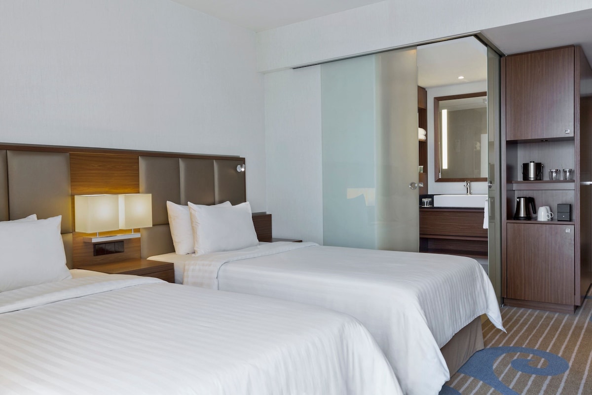 A hotel room with two beds, a nightstand with a lamp, a wooden headboard, a glass partition, a bathroom with a mirror, and a kitchenette with a coffee maker.